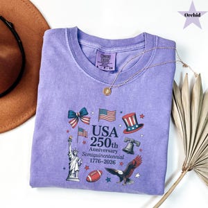 May include: A lilac-colored t-shirt featuring a graphic commemorating the USA's 250th anniversary. The design includes American flags, the Statue of Liberty, an eagle, and the text "USA 250th Anniversary Semiquincentennial 1776-2026". A gold necklace rests on the shirt.