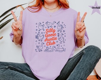 May include: Orchid-colored t-shirt with a graphic of cartoon geese and the text "Silly Goose Auntie Club." The design includes hearts, stars, and floral elements. The shirt is a relaxed fit.