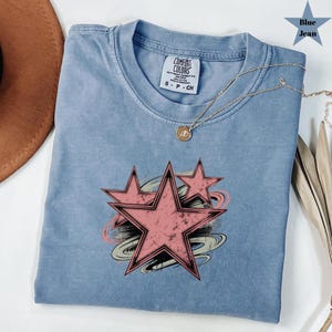 May include: A blue jean Comfort Colors t-shirt with a distressed pink star graphic. The shirt has a crew neck and short sleeves. A gold necklace with a pendant is draped across the shirt.