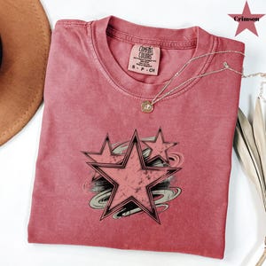 May include: A dusty rose Comfort Colors t-shirt featuring a distressed star graphic with black outlines and gray accents. The shirt has a crew neck and short sleeves. A silver necklace with a small pendant is draped across the shirt.