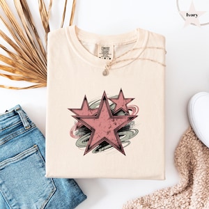 May include: Ivory-colored t-shirt featuring a distressed pink star design with a swirling gray and pink background. The shirt has a crew neck and short sleeves. The Comfort Colors tag is visible. The shirt is folded on a white surface.