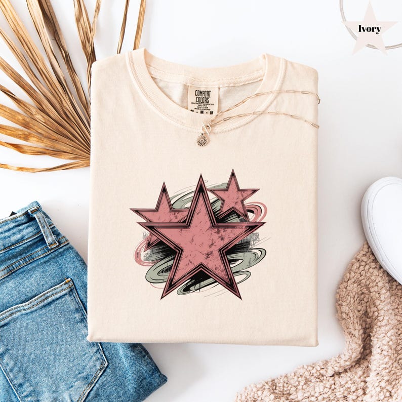 May include: Ivory-colored t-shirt featuring a distressed pink star design with a swirling gray and pink background. The shirt has a crew neck and short sleeves. The Comfort Colors tag is visible. The shirt is folded on a white surface.