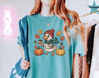May include: Teal t-shirt featuring a cartoon duck wearing a red beanie and scarf, holding a coffee cup, and walking between two pumpkins. Autumn leaves and acorns surround the duck, creating a fall theme.