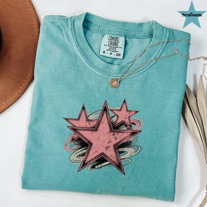 May include: Seafoam green t-shirt with a distressed pink star graphic. The shirt has a crew neck and short sleeves. A gold necklace with a pendant is draped over the shirt. The tag reads "Comfort Colors".