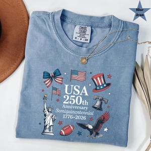 May include: Blue jean Comfort Colors t-shirt with a graphic celebrating the USA's 250th Anniversary. The design includes the Statue of Liberty, American flags, a top hat, a football, and an eagle. A gold necklace is draped across the shirt.