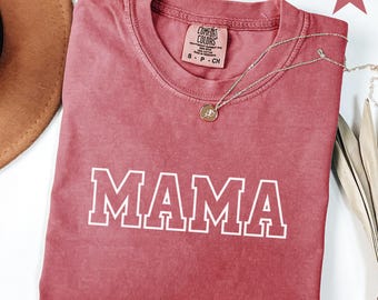 May include: A crimson-colored t-shirt with the word "MAMA" printed in white block letters. The shirt has a crew neck and a small tag near the collar. A silver necklace with a round pendant is draped across the shirt.