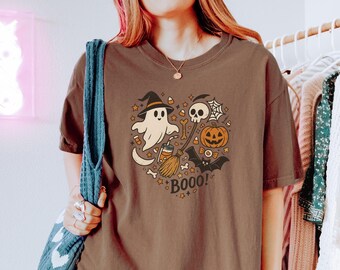 May include: Brown t-shirt with a Halloween-themed graphic. The design features a ghost wearing a witch hat, a skull, a pumpkin, a bat, and the word "BOOO!" in a playful font. The graphic is surrounded by stars and other Halloween elements.