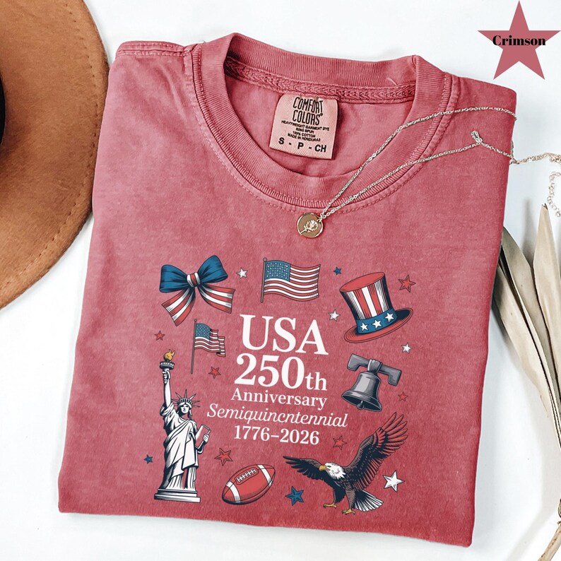 May include: Crimson red t-shirt with a graphic commemorating the USA's 250th anniversary. The design includes the Statue of Liberty, American flags, a football, an eagle, and the text "USA 250th Anniversary Semiquincentennial 1776-2026".