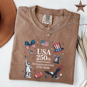 May include: Espresso-colored t-shirt with a graphic celebrating the USA's 250th anniversary. The design includes American flags, a bow, a top hat, the Liberty Bell, an eagle, and the Statue of Liberty. The text reads "USA 250th Anniversary Semiquincentennial 1776-2026."