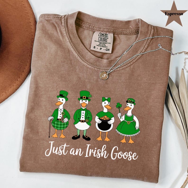 Puede incluir: Camiseta color espresso con un gr&aacute;fico de cuatro gansos de dibujos animados vestidos para el D&iacute;a de San Patricio. Los gansos llevan sombreros, chaquetas y vestidos verdes, y sostienen un bast&oacute;n, una olla de oro y un tr&eacute;bol. El texto "Just an Irish Goose" est&aacute; impreso debajo.