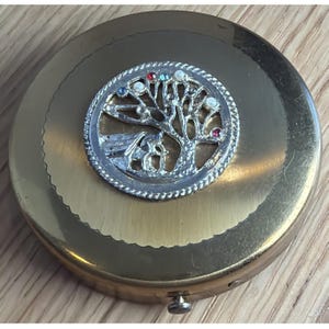 May include: A round, gold-toned compact with a decorative silver-colored emblem. The emblem features a tree design with colorful gemstones and a small house. The compact has a hinged opening and a textured edge.