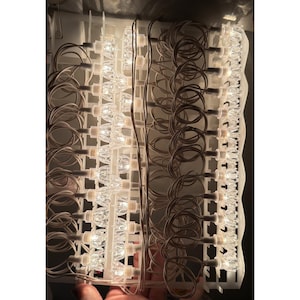 May include: A set of clear, faceted, diamond-shaped light bulbs arranged in vertical rows on a white plastic grid. The bulbs are connected by thin white wires, creating a structured display of multiple small, glowing, decorative lights.