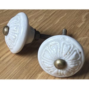 May include: Two white ceramic cabinet knobs with a floral design and brass-colored hardware. The knobs are set against a wooden surface, showcasing their decorative details and vintage style. The knobs are approximately 1.5 inches in diameter.