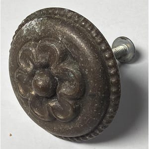 May include: A bronze cabinet knob featuring a central floral motif. The knob is circular, with a beaded border and a silver screw. The finish has a slightly aged appearance. The knob's diameter is approximately 4 cm.