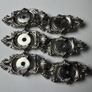 May include: Six silver-coloured, ornate drawer pulls. Each pull features a central circular area with a hole, surrounded by an elaborate, symmetrical scrollwork design. The pulls are arranged in two rows of three.