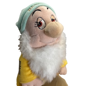 May include: A plush doll of the character Dopey from Snow White. The doll has a light green hat, a large white beard, and a yellow shirt. The doll's face is detailed with embroidered eyes and a large nose.