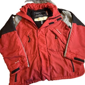 Puede incluir: Chaqueta de invierno Polo Jeans Co. roja y negra. La chaqueta tiene capucha, múltiples bolsillos y el texto "RL-67" en la manga. La chaqueta tiene cremallera negra y cuello negro.