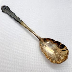 May include: A vintage silver spoon with a decorative, scalloped bowl and handle. The handle has a textured, dark gray finish, while the bowl and stem are a lighter silver color. The spoon is designed for serving.