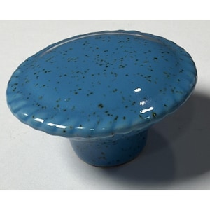 May include: A blue ceramic cabinet knob with a speckled surface and a scalloped edge. The knob has a rounded top and a short, cylindrical base. The color is a vibrant, glossy blue with darker speckles.