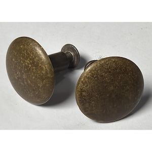 May include: Two antique brass cabinet knobs with a distressed finish. Each knob has a rounded, oval face and a short, cylindrical stem. The knobs are a warm, aged brass color, suggesting vintage hardware for furniture restoration or home decor.