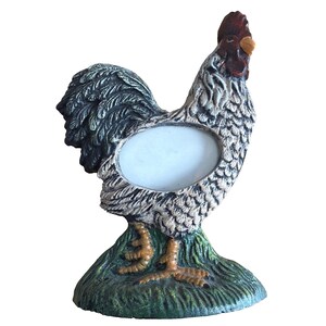 May include: A rooster-shaped photo frame. The frame is painted in shades of gray, brown, and green, with a red comb and beak. The frame has an oval opening for a photo and sits on a green base.