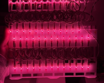 Puede incluir: Una tira de bombillas incandescentes en miniatura que emiten un brillo rosa intenso, organizadas en filas sobre un marco de almacenamiento de plástico blanco. Las bombillas están conectadas por cables blancos finos enrollados entre cada fila.
