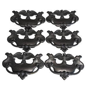 Pode incluir: Seis puxadores de gaveta em estilo antigo em metal escuro, cada um com um desenho de águia. Os puxadores têm uma pega curva e uma placa traseira ornamentada. A águia está centrada acima da pega, com as asas abertas.