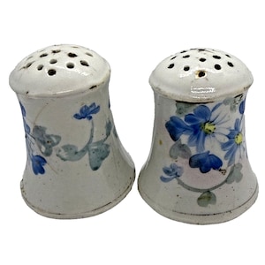 May include: A pair of white ceramic salt and pepper shakers. Each shaker features a flared base and a domed top with multiple holes. The shakers are decorated with blue floral designs and green leaves. The overall style is vintage.