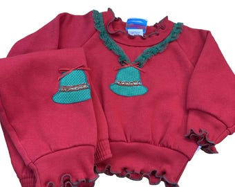 Op de afbeelding: Een rood kerstoutfit bestaande uit een top en broek. De top en broek hebben een groene bel-appliqué met een strik en decoratieve rand. De halslijn en manchetten hebben groene ruches. De outfit is ontworpen voor kinderen.