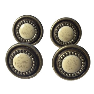 Puede incluir: Cuatro tiradores de armario de estilo antiguo. Cada tirador presenta un centro color crema con detalles moteados, rodeado por un anillo de pequeñas cuentas en relieve. El anillo exterior es de color bronce oscuro envejecido.