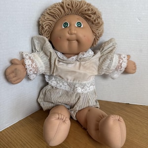 May include: A vintage Cabbage Patch Kid doll with a tan complexion, green eyes, and a soft, yarn-like hairstyle. The doll is dressed in a cream-coloured outfit with lace trim and is seated with its arms and legs extended.