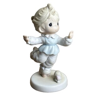 May include: A porcelain figurine of a child in light blue pajamas with white polka dots, balancing on one foot. The figurine has a round base and a small, matching figurine of a turtle is next to the child.