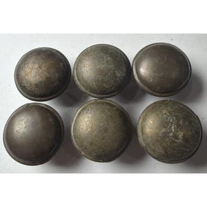 Puede incluir: Seis tiradores de armario antiguos de color bronce, con forma redondeada y abovedada. Los tiradores tienen un aspecto envejecido, con variaciones de color y textura, lo que sugiere edad y uso. Están dispuestos en dos filas de tres.