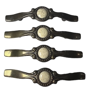 May include: Four antique bronze cabinet pulls with a cream-colored, speckled center. The pulls have a decorative, curved design with a round center and are likely for drawers or cabinets. The hardware has a vintage aesthetic.