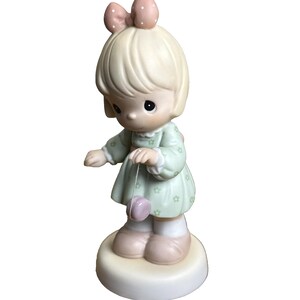 May include: A ceramic figurine of a young girl in a mint green dress with a pink bow and a matching yo-yo. The figurine has a light skin tone and is standing on a white base.
