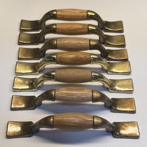 May include: A set of seven cabinet handles arranged in a vertical row. Each handle features a central cylindrical light brown wood grip flanked by two curved metallic brass-toned ends with flat rectangular mounting plates at the outer edges.