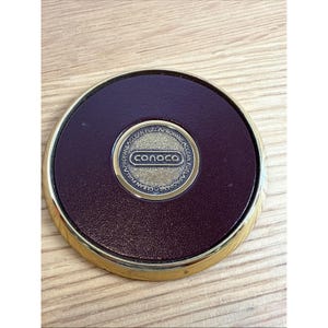 May include: A round, maroon-colored Conoco fuel cap with a gold-colored rim. The center features the Conoco logo in a gold oval, surrounded by the words "Propane Clean Fuel".