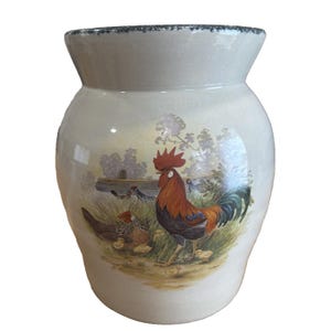 May include: A ceramic vase with a cream-colored body and a flared top. The vase features a painted scene of a rooster, hen, and chicks in a grassy field. The rooster has red, orange, and black feathers. The top rim has a dark blue trim.