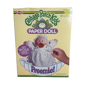 Pode incluir: Uma caixa de boneca de papel Cabbage Patch Kids vintage. A caixa apresenta uma boneca com uma chupeta e a palavra "Preemie!". A caixa também inclui o texto "Paper Doll" e "Ages: 4 thru 8".