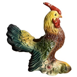 May include: A vintage ceramic rooster planter in shades of yellow, red, green, and brown. The rooster has a detailed design with a red comb and wattle. The planter is designed to hold plants or flowers.