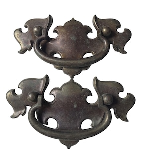 Pode incluir: Duas puxadores de gaveta antigos em bronze. Cada puxador apresenta um design decorativo e ornamentado com uma alça curva e detalhes florais estilizados. Os puxadores são feitos de metal e têm um acabamento desgastado, sugerindo idade e uso.