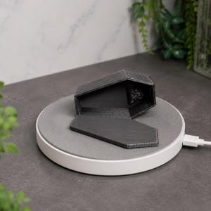 May include: A black 3D-printed coffin-shaped container with a removable lid, resting on a circular grey wireless charging pad with a white base and a connected USB cable extending from the side of the device.