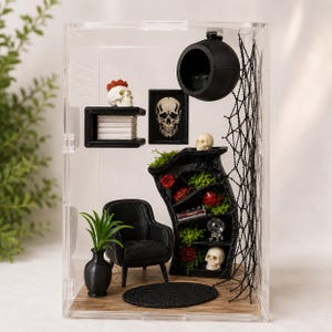 Puede incluir: Una habitaci&oacute;n en miniatura dentro de una caja de acr&iacute;lico transparente con un sill&oacute;n negro, una alfombra redonda negra y una estanter&iacute;a negra curva con rosas rojas, musgo verde y calaveras. Una esfera negra cuelga con una red y calaveras.