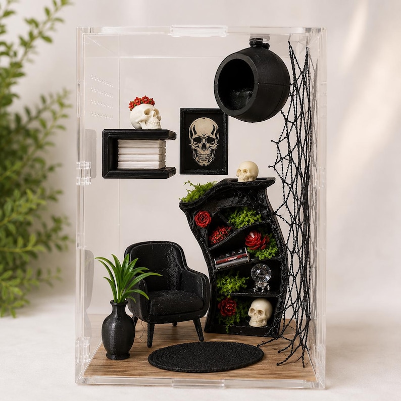 Puede incluir: Una habitaci&oacute;n en miniatura dentro de una caja de acr&iacute;lico transparente con un sill&oacute;n negro, una alfombra redonda negra y una estanter&iacute;a negra curva con rosas rojas, musgo verde y calaveras. Una esfera negra cuelga con una red y calaveras.