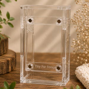 Puede incluir: Un terrario rectangular transparente de acrílico con el texto "Tiny Fur Forage" grabado en la parte delantera. El recinto tiene pequeños orificios de ventilación y está sujeto con cuatro remaches de color marrón oscuro. El terrario se exhibe sobre una superficie de madera.