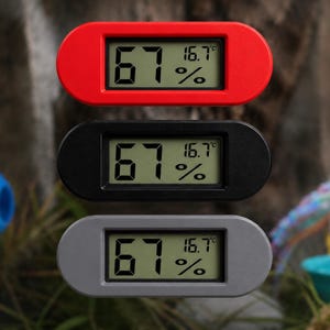 May include: Three digital hygrometers with rounded rectangular frames in red, black, and grey. Each device features a central LCD screen displaying 67% humidity and a temperature of 16.7 degrees Celsius in large, clear black digital numerals.