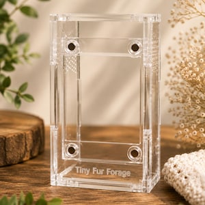 Puede incluir: Un terrario rectangular transparente de acrílico con el texto "Tiny Fur Forage" grabado en la parte delantera. El terrario tiene una abertura rectangular y pequeños orificios de ventilación. El fondo incluye vegetación y una superficie de madera.