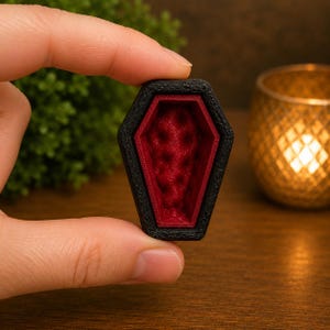 May include: A small, coffin-shaped object with a textured black exterior frame and a deep red interior featuring a tufted, padded-style pattern. The item has a hexagonal silhouette with a pointed top and bottom, showcasing a stark contrast between the dark outer border and the vibrant, recessed crimson centre.