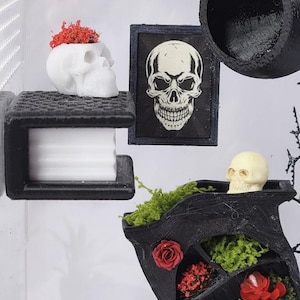 Puede incluir: Una estanter&iacute;a negra en miniatura con musgo verde y flores rojas, una maceta blanca con forma de cr&aacute;neo con flores rojas, una impresi&oacute;n de cr&aacute;neo enmarcada y un peque&ntilde;o cr&aacute;neo beige. Una estructura negra impresa en 3D sostiene un bloque blanco.