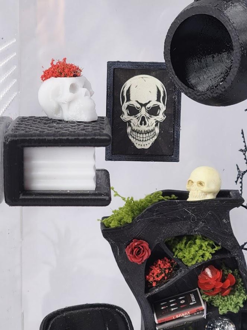 Puede incluir: Una estanter&iacute;a negra en miniatura con musgo verde y flores rojas, una maceta blanca con forma de cr&aacute;neo con flores rojas, una impresi&oacute;n de cr&aacute;neo enmarcada y un peque&ntilde;o cr&aacute;neo beige. Una estructura negra impresa en 3D sostiene un bloque blanco.